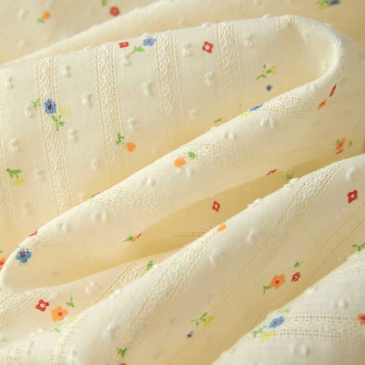 Gros plan sur le Tissu plumetis à petites fleurs au mètre de Longan Craft France, un coton plumetis doux de couleur crème avec des points texturés et de petits imprimés floraux rouges, orange et bleus, idéal pour les vêtements confortables.