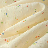 Gros plan sur le Tissu plumetis à petites fleurs au mètre de Longan Craft France, un coton plumetis doux de couleur crème avec des points texturés et de petits imprimés floraux rouges, orange et bleus, idéal pour les vêtements confortables.