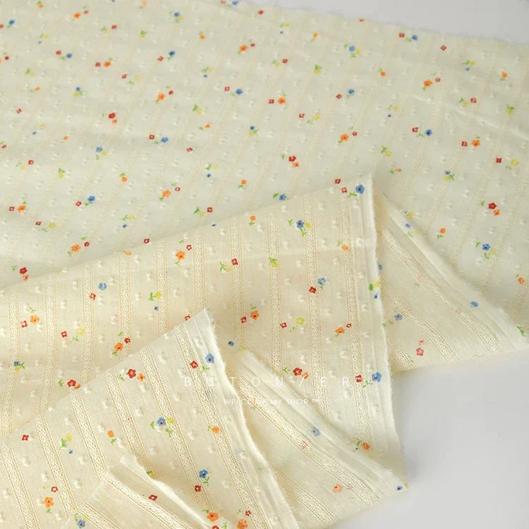 Un morceau plié du tissu plumetis en coton crème doux de Longan Craft France, qui présente une texture rayée subtile et de petits imprimés floraux dispersés - idéal pour des vêtements confortables.