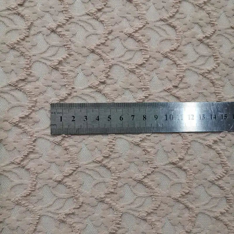 Une règle argentée mesurant les centimètres est posée sur la Dentelle extensible au mètre de Longan Craft France, une dentelle extensible beige clair brodée de franges florales, mettant en valeur la texture et le design détaillés du tissu.