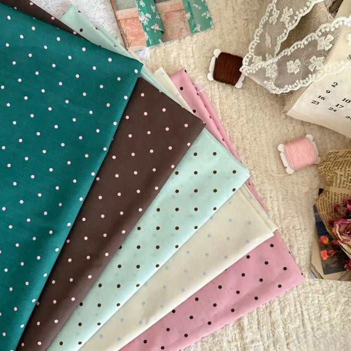 Six pièces de linon de coton imprimé à petits pois Longan Craft France, en teal, marron, menthe, bleu clair, crème et rose, sont disposées avec dentelle et bobines sur une surface texturée pour créations couture décontractées.