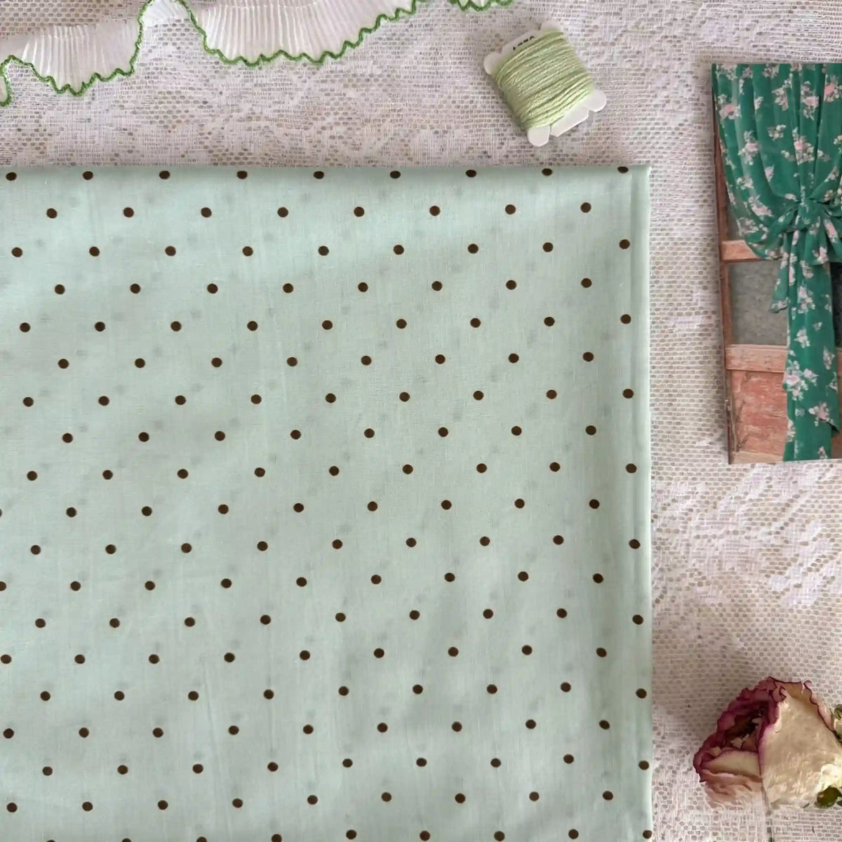 Un morceau de Linon de coton imprimé à petits pois de Longan Craft France est posé sur de la dentelle, avec une bobine de fil vert, une bordure de rickrack vert, une photo de rideau floral et une rose séchée à proximité.