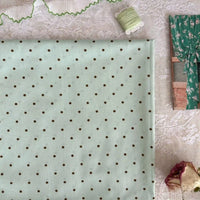 Un morceau de Linon de coton imprimé à petits pois de Longan Craft France est posé sur de la dentelle, avec une bobine de fil vert, une bordure de rickrack vert, une photo de rideau floral et une rose séchée à proximité.