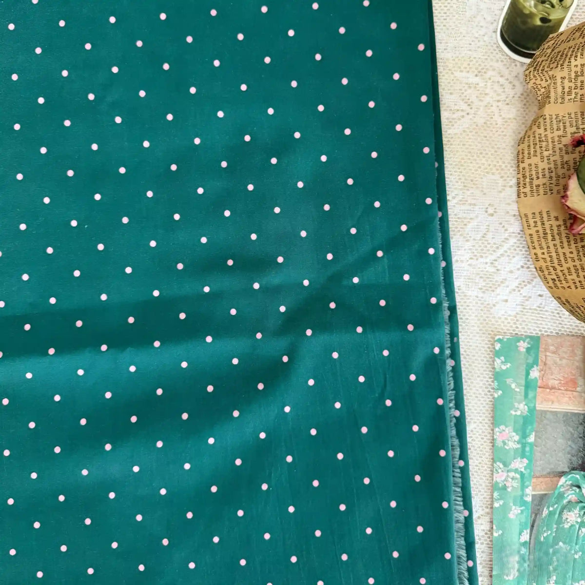 Un morceau de Linon de coton imprimé à petits pois au mètre par Longan Craft France est présenté sur de la dentelle blanche, à côté d'un panier contenant du papier imprimé, des fleurs séchées et des feuilles de papier à motifs. Ce tissu 100 % coton est léger et respirant, idéal pour les tenues décontractées.