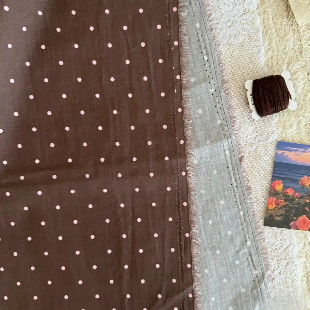 Un morceau de Linon de coton imprimé à petits pois au mètre par Longan Craft France est posé sur une surface blanche texturée, à côté d'une bobine de fil sombre et d'une carte postale représentant des fleurs et un coucher de soleil.