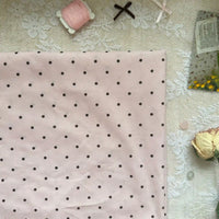 Un morceau de Linon de coton imprimé à petits pois au mètre par Longan Craft France est posé sur de la dentelle, avec une bobine de fil rose, un ruban, une fleur séchée et un sachet floral à proximité. Ce tissu linon 100% coton léger est parfait pour une tenue décontractée.