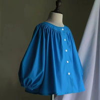 Un mannequin présente la "Blouse à encolure ronde smockée avec manches raglan" de Longan Craft France, un haut bleu ample avec des manches froncées et des boutons blancs. Fond uni, tissu blanc drapé sur la droite. Patron de couture en PDF à télécharger gratuitement.