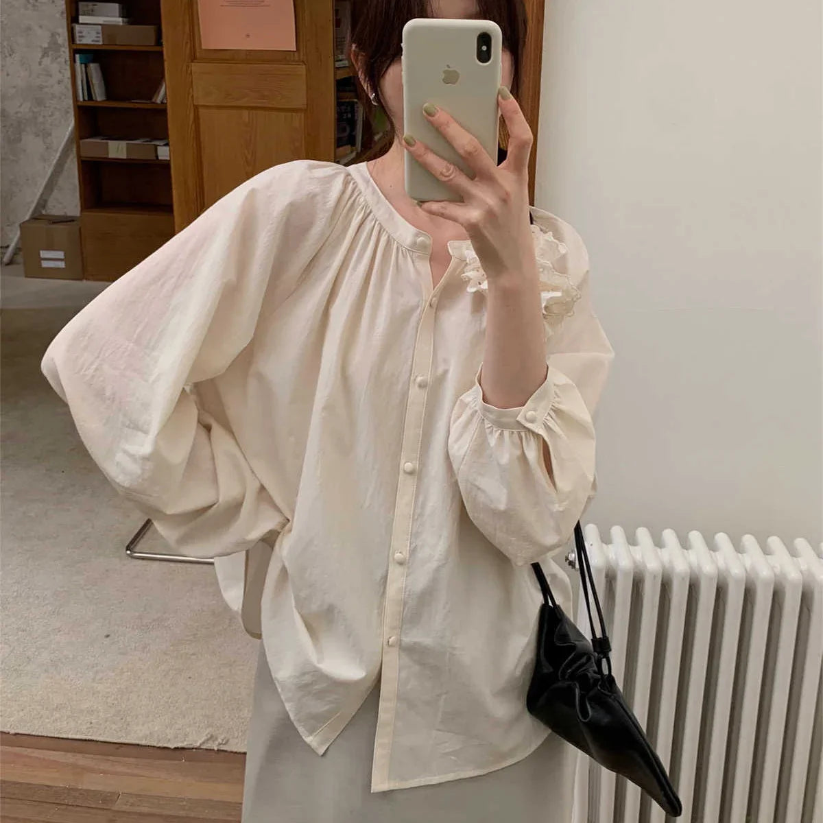 Une personne se tient à l'intérieur et prend un selfie miroir avec un smartphone. Elle porte le Longan Craft France Blouse à encolure ronde smockée avec manches raglan (patron de couture gratuit en PDF) et tient un petit sac noir. Des étagères, des boîtes et un radiateur sont à l'arrière-plan.