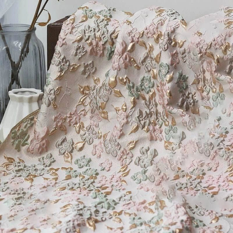 Le Tissu brocart floral à bulles douces de Longan Craft France, avec sa base rose clair ornée de motifs fleuris pastel, apporte une texture unique à la déco. À l'arrière, un vase transparent et un vase blanc subliment l'ensemble.