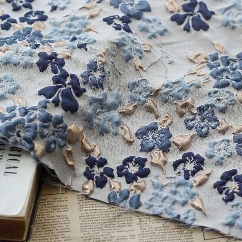 Le Longan Craft France Tissu brocart floral à bulles douces au mètre, orné de broderies florales bleues et beiges, est drapé sur des livres et journaux anciens, ajoutant une touche texturée unique qui rehausse n'importe quel décor.