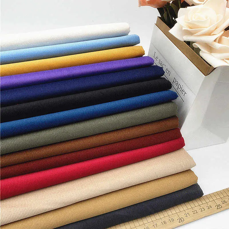 Des piles de Tissu velours côtelé uni 21 côtes au mètre de Longan Craft France, dans des couleurs assorties, sont présentées à côté d'une boîte blanche contenant des roses artificielles, avec une règle à la base pour montrer ce tissu de coton confortable pour les vêtements.