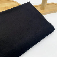 Une pièce de Longan Craft France Tissu velours côtelé uni 21 côtes au mètre, en coton noir à fines côtes, est pliée sur un plateau en bois léger, avec son bord relevé visible. Ce tissu confortable est parfait pour confectionner des pantalons élégants ou d'autres vêtements.