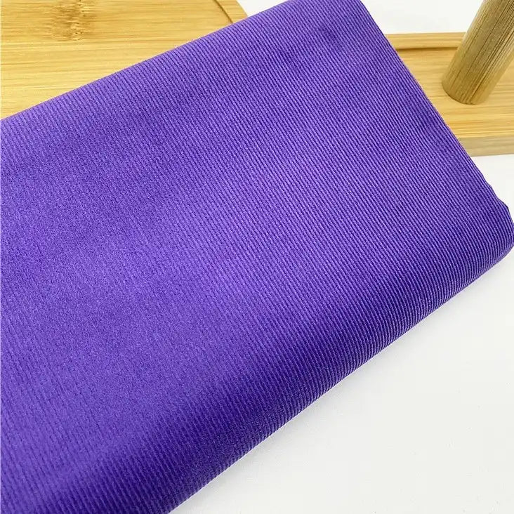 Gros plan sur le tissu velours côtelé uni 21 côtes au mètre de Longan Craft France, un tissu velours côtelé en coton doux avec de fines nervures, plié sur une surface en bois clair avec une partie d'un objet en bois à l'arrière-plan.