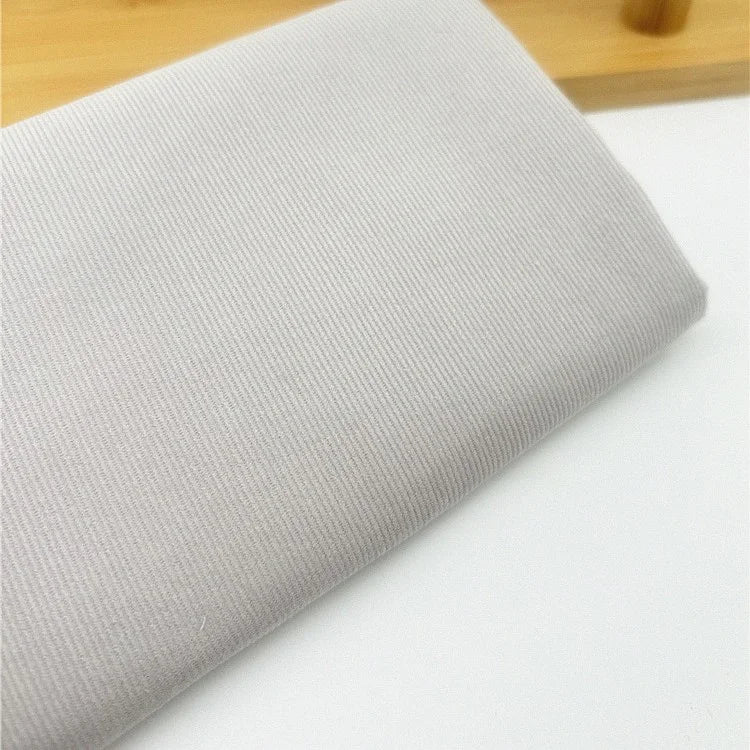 Un gros plan du Tissu velours côtelé uni 21 côtes au mètre de Longan Craft France montre le tissu velours côtelé de coton gris clair avec de fines nervures plié sur une surface blanche, avec une partie d'un objet en bois à l'arrière-plan.