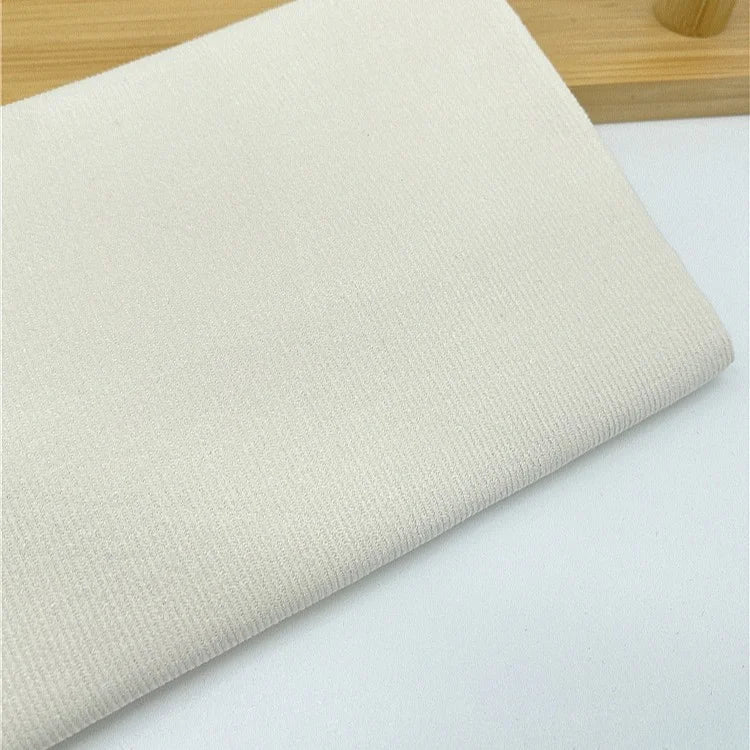 Gros plan d'un pli Longan Craft France Tissu velours côtelé uni 21 côtes au mètre, un velours côtelé en coton à fines côtes pour vêtements, sur une surface blanche avec un objet en bois clair en arrière-plan.