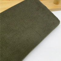 Gros plan sur le Tissu velours côtelé uni 21 côtes au mètre en coton, coloris olive, de Longan Craft France, plié sur une surface claire à côté d'un objet en bois. Idéal pour vêtements confortables.