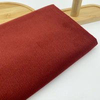 Gros plan sur le Tissu velours côtelé uni 21 côtes au mètre en coton de Longan Craft France, de couleur rouille, présenté sur une surface en bois clair avec un support en bois en arrière-plan. Parfait pour les projets de vêtements confortables en velours côtelé.