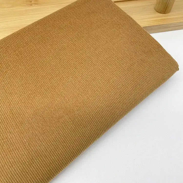 Gros plan sur le tissu velours côtelé uni 21 côtes au mètre de Longan Craft France, un velours côtelé de coton marron clair à fines côtes destiné à l'habillement, plié sur une surface blanche avec une partie d'une structure en bois à l'arrière-plan.