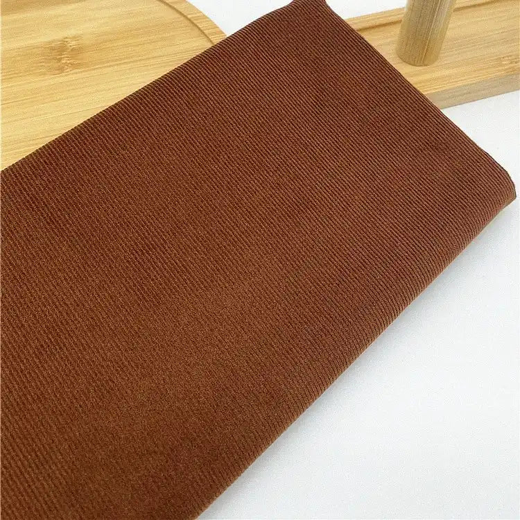 Un morceau de tissu Longan Craft France velours côtelé uni 21 côtes en coton, idéal pour vêtements, est plié sur une surface en bois clair avec un objet en bois visible à l'arrière-plan.