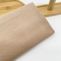 Gros plan sur le Tissu velours côtelé uni 21 côtes au mètre de Longan Craft France en beige clair, plié sur une surface blanche avec une partie d'un plateau en bois et une tige verticale en arrière-plan.