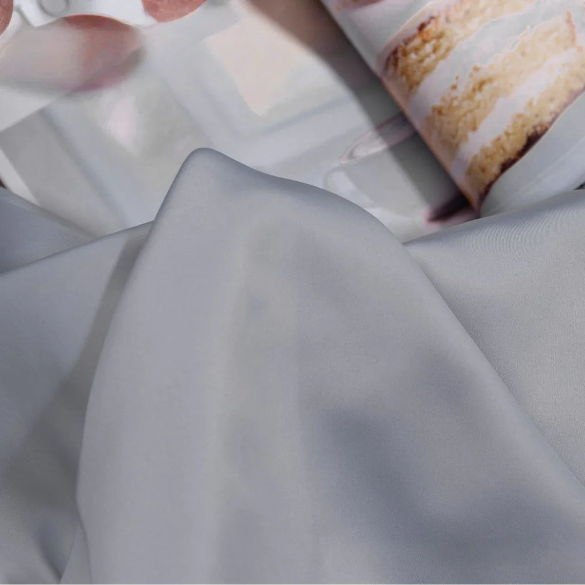 Gros plan sur le Tissu satin charmeuse uni au mètre de Longan Craft France, un crêpe épais de satin gris clair lisse pour les robes de mariée ou de soirée, recouvrant partiellement une page de magazine avec un gâteau de couches givré blanc.