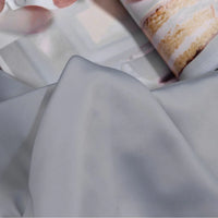 Gros plan sur le Tissu satin charmeuse uni au mètre de Longan Craft France, un crêpe épais de satin gris clair lisse pour les robes de mariée ou de soirée, recouvrant partiellement une page de magazine avec un gâteau de couches givré blanc.