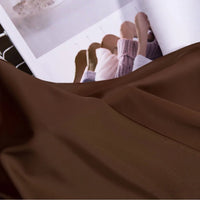 Un morceau de Tissu satin charmeuse uni au mètre de Longan Craft France est drapé au premier plan, tandis qu'un magazine ouvert à l'arrière-plan présente des images de pulls sur des cintres en bois.