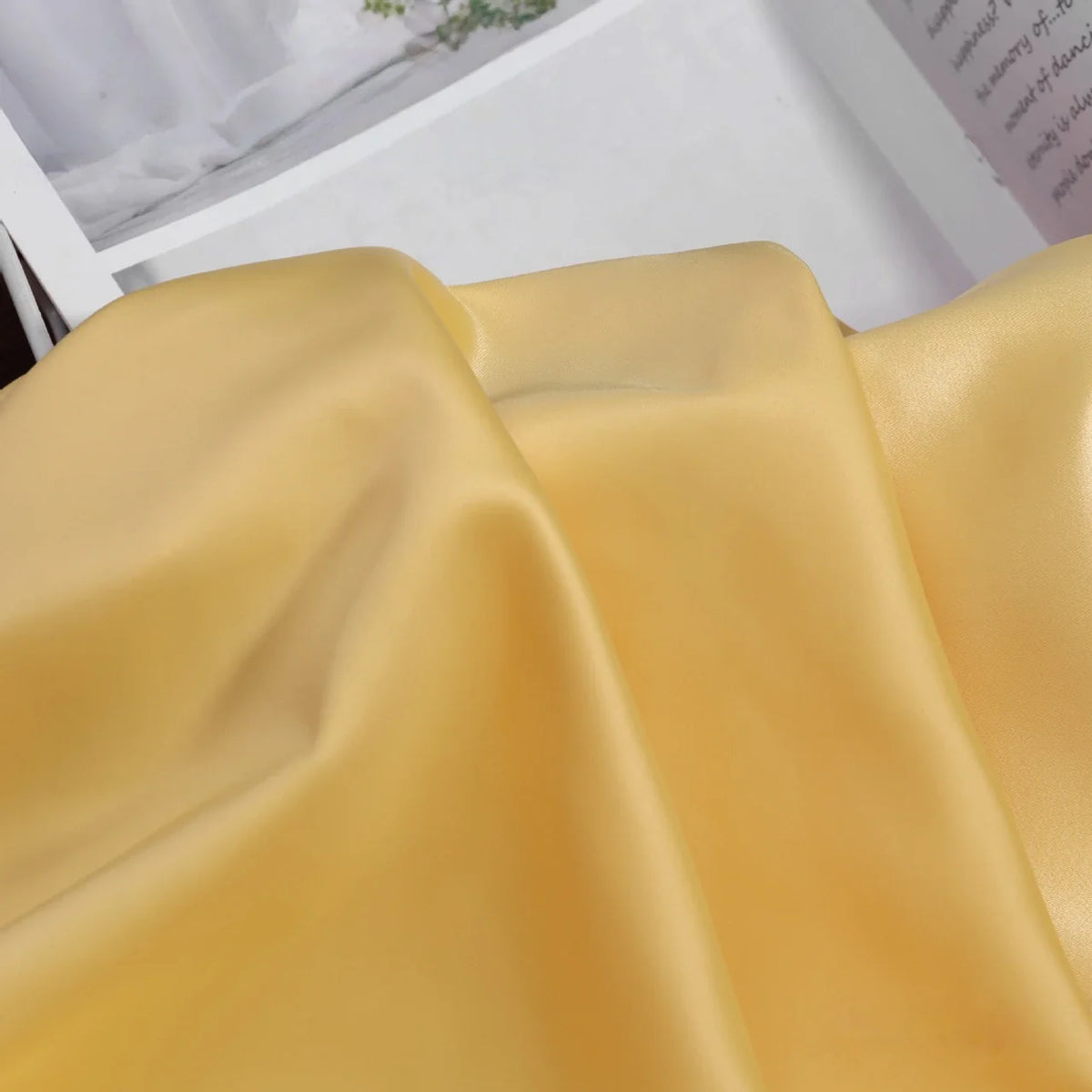 Gros plan sur le tissu satin charmeuse uni au mètre de Longan Craft France, un crêpe épais lisse en satin jaune parfait pour les robes de mariée ou de soirée, drapé sur une surface avec un magazine ouvert visible à l'arrière-plan.