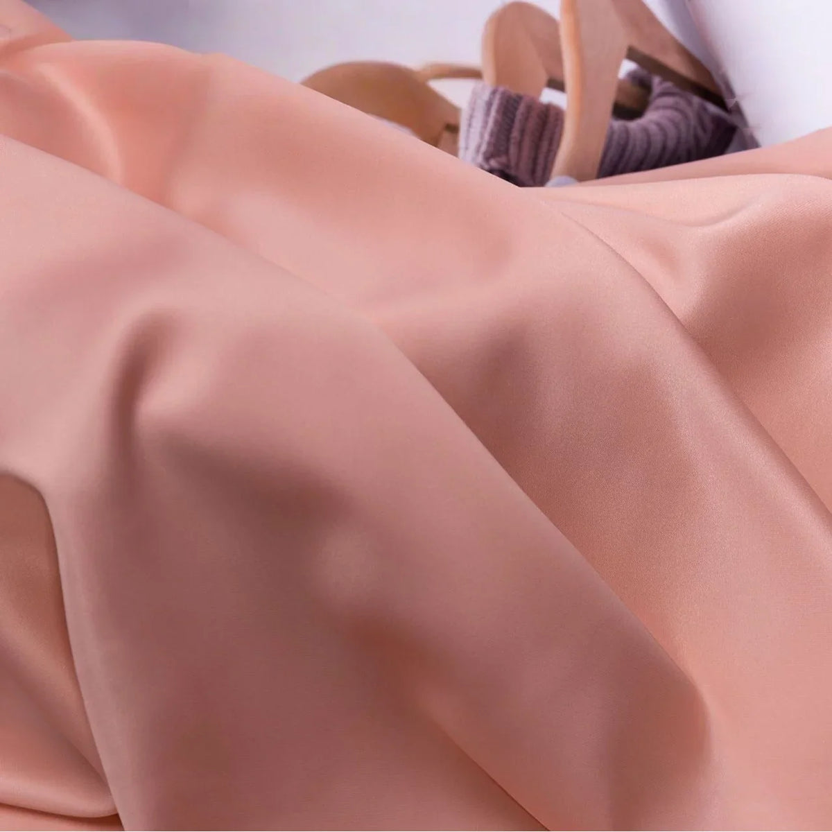 Gros plan sur le tissu satin charmeuse uni au mètre de Longan Craft France, de couleur pêche avec des plis subtils. En arrière-plan, des sandales à talons de couleur bronze et un article côtelé de couleur mauve - idéal pour robes de mariée ou de soirée élégantes.