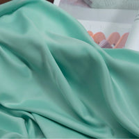 Gros plan sur le tissu satin charmeuse uni de Longan Craft France en vert menthe clair, avec une texture lisse et soyeuse et des plis doux. En arrière-plan, des couleurs pastel floues. Idéal pour robes de mariée et robes de soirée.