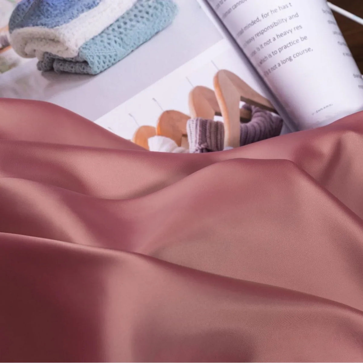 Gros plan sur le Tissu satin charmeuse uni au mètre de Longan Craft France, drapé devant un magazine ouvert avec des tricots pliés et des vêtements suspendus, mettant en valeur ce crêpe satin épais pour robes de mariée et robes de soirée.