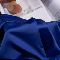 Gros plan sur le tissu satin charmeuse uni au mètre de Longan Craft France, un crêpe épais de satin bleu brillant destiné aux robes de mariée ou de soirée. Il est librement drapé devant un magazine ouvert présentant des images de plantes.