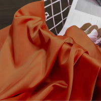 Gros plan sur le Tissu satin charmeuse uni au mètre de Longan Craft France, un crêpe épais de satin orange brillant pour les robes de mariée ou de soirée, avec des plis souples sur une surface sombre, à côté d'un magazine de chaussures et d'un objet filaire blanc.