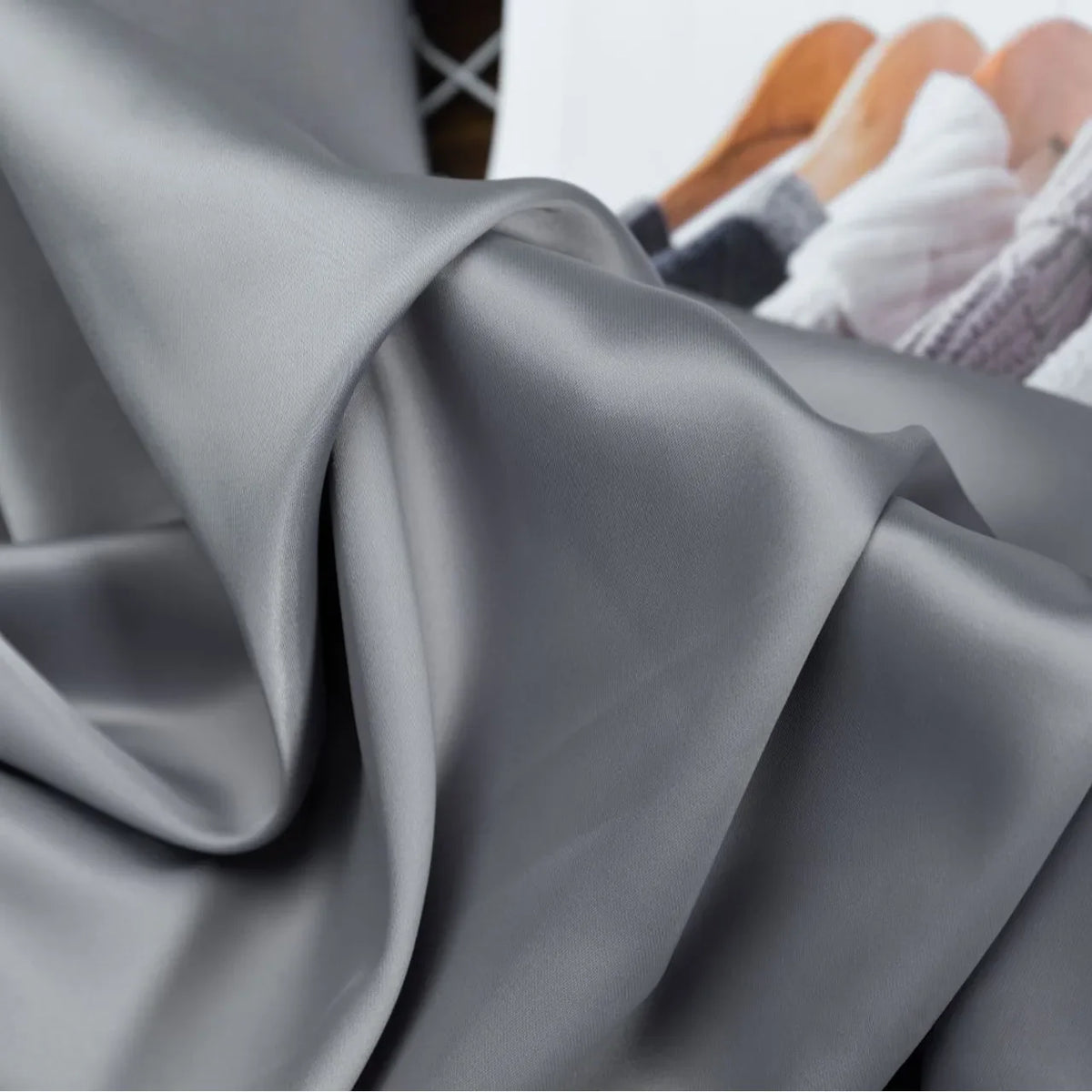 Tissu satin charmeuse uni au mètre de Longan Craft France, lisse et gris argenté, drapé en plis souples, avec un arrière-plan flou représentant une partie d'une étagère à vêtements et des vêtements de couleur claire. Un crêpe épais en satin parfait pour les robes de mariée ou de soirée.