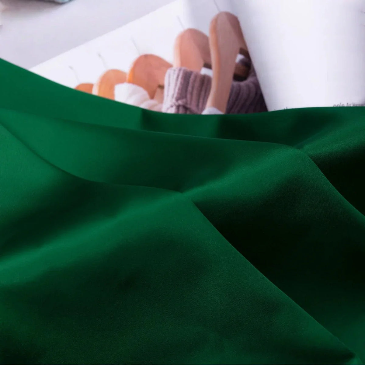 Au premier plan, un morceau de Tissu satin charmeuse uni au mètre de Longan Craft France est drapé, tandis qu'à l'arrière-plan, un magazine ouvert présente des cintres en bois avec des tricots.