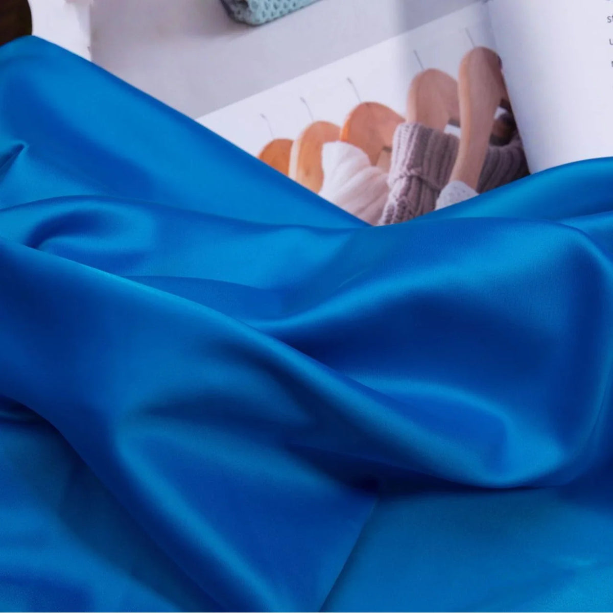 Un morceau de Tissu satin charmeuse uni de Longan Craft France est posé devant un magazine ouvert présentant des tricots sur des cintres en bois. Ce textile épais en crêpe de satin est idéal pour les robes de mariée ou de soirée.