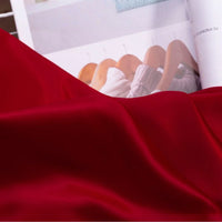 Un gros plan du Tissu satin charmeuse uni au mètre de Longan Craft France - un crêpe épais de satin rouge et lisse pour les robes de mariée et de soirée - recouvre en partie un magazine ouvert avec des cintres en bois et des vêtements légers.