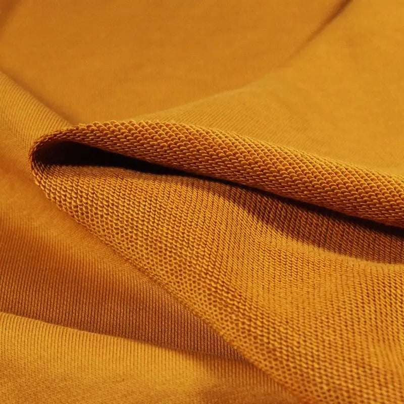Gros plan sur le tissu Longan Craft France Tissu French Terry en coton uni au mètre en jaune moutarde, montrant le tissage et la texture du tissu avec un bord plié révélant à la fois ses côtés rugueux et lisses - idéal pour les sweatshirts et les sweat-shirts à capuche.