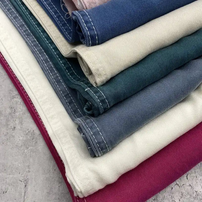 Une pile de tissu denim lyocell multicolore de Longan Craft France (vert, bleu, crème, violet et rose) présente des coutures blanches visibles. Ce textile respirant et durable pour les jeans est présenté sur une surface texturée grise.