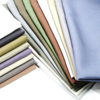 Une pile de tissu lyocell et lin uni au mètre de Longan Craft France dans les coloris bleu, vert, marron, violet, blanc et beige est soigneusement superposée sur un fond blanc. Ce textile durable et infroissable est parfait pour les peignoirs respirants.