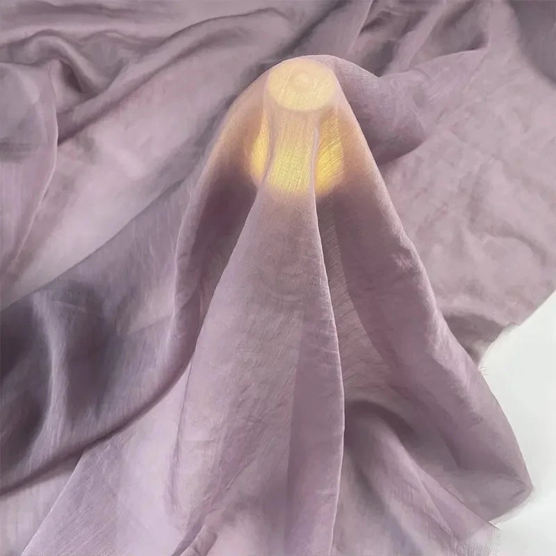 Une petite source lumineuse ronde est recouverte de Tissu lin et soie uni au mètre de Longan Craft France, un mélange haut de gamme de lin, de lyocell et de soie pour l'ameublement, qui projette une douce lueur lavande à travers le tissu luxueux drapé en douceur sur une surface.