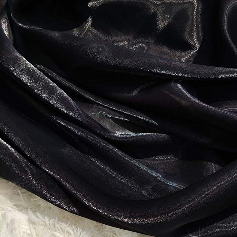 Gros plan sur l'organza noir et brillant de Longan Craft France, drapé en plis lâches et reflétant la lumière. Le tissu organza lisse et transparent uni au mètre contraste avec une surface texturée blanche dans le coin inférieur gauche.