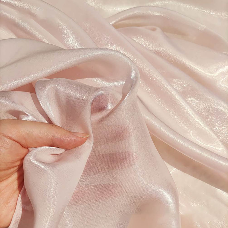 Une main présente le Tissu organza uni au mètre de Longan Craft France, un tissu léger et transparent idéal pour vêtements et décoration, en rose pâle avec une finition brillante et réfléchissante sur le tissu assorti.