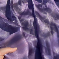 Une main tient un uni au mètre en Tissu organza brillant, violet et semi-transparent de Longan Craft France, dont les plis délicats reflètent la lumière - idéal pour l'habillement et la décoration.
