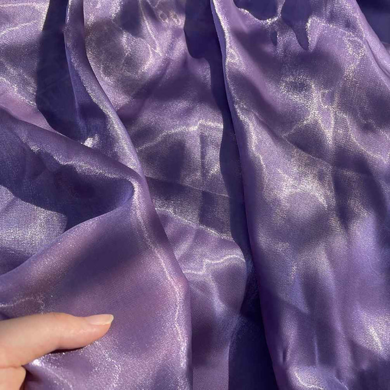 Une main tient un uni au mètre en Tissu organza brillant, violet et semi-transparent de Longan Craft France, dont les plis délicats reflètent la lumière - idéal pour l'habillement et la décoration.