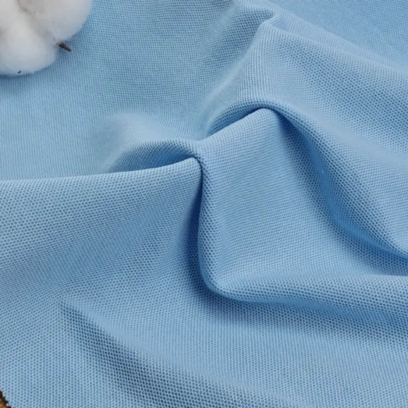 Un tissu bleu clair piqué uni au mètre de Longan Craft France aux plis souples et au tissage visible est présenté, avec un petit morceau de tissu blanc en coton-polyester visible dans le coin supérieur gauche. Idéal pour bricoler des chemises et des polos d'été.
