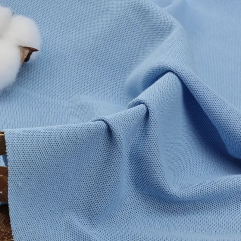 Un gros plan du tissu piqué uni au mètre de Longan Craft France montre sa maille texturée bleu clair en coton-polyester - un tissu idéal pour les polos et les chemises à manches courtes de l'été. Le détail en coton en haut à gauche souligne sa qualité spécifique pour les polos.
