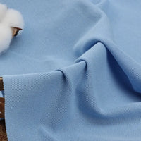 Un gros plan du tissu piqué uni au mètre de Longan Craft France montre sa maille texturée bleu clair en coton-polyester - un tissu idéal pour les polos et les chemises à manches courtes de l'été. Le détail en coton en haut à gauche souligne sa qualité spécifique pour les polos.