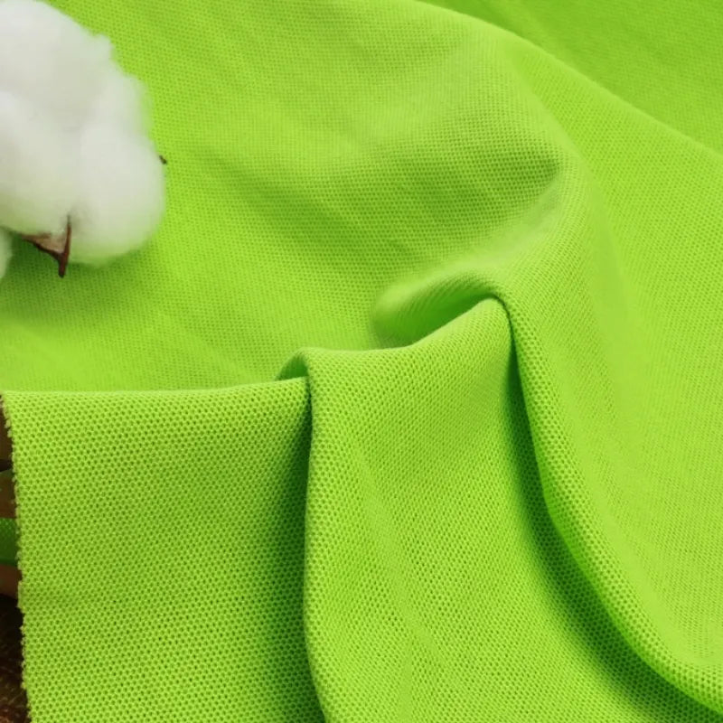 Un gros plan montre le tissu piqué uni au mètre de Longan Craft France, un tissu vert vif en coton-polyester pour les chemises de bricolage et les polos d'été, drapé en plis souples avec du coton blanc et une tige brune dans le coin supérieur gauche.