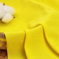 Gros plan sur un tissu piqué uni au mètre jaune vif de Longan Craft France, dont la surface texturée en coton-polyester est drapée sur un panier tressé. Un petit morceau de coton brut apparaît sur la gauche. Idéal pour les chemises et les polos d'été.